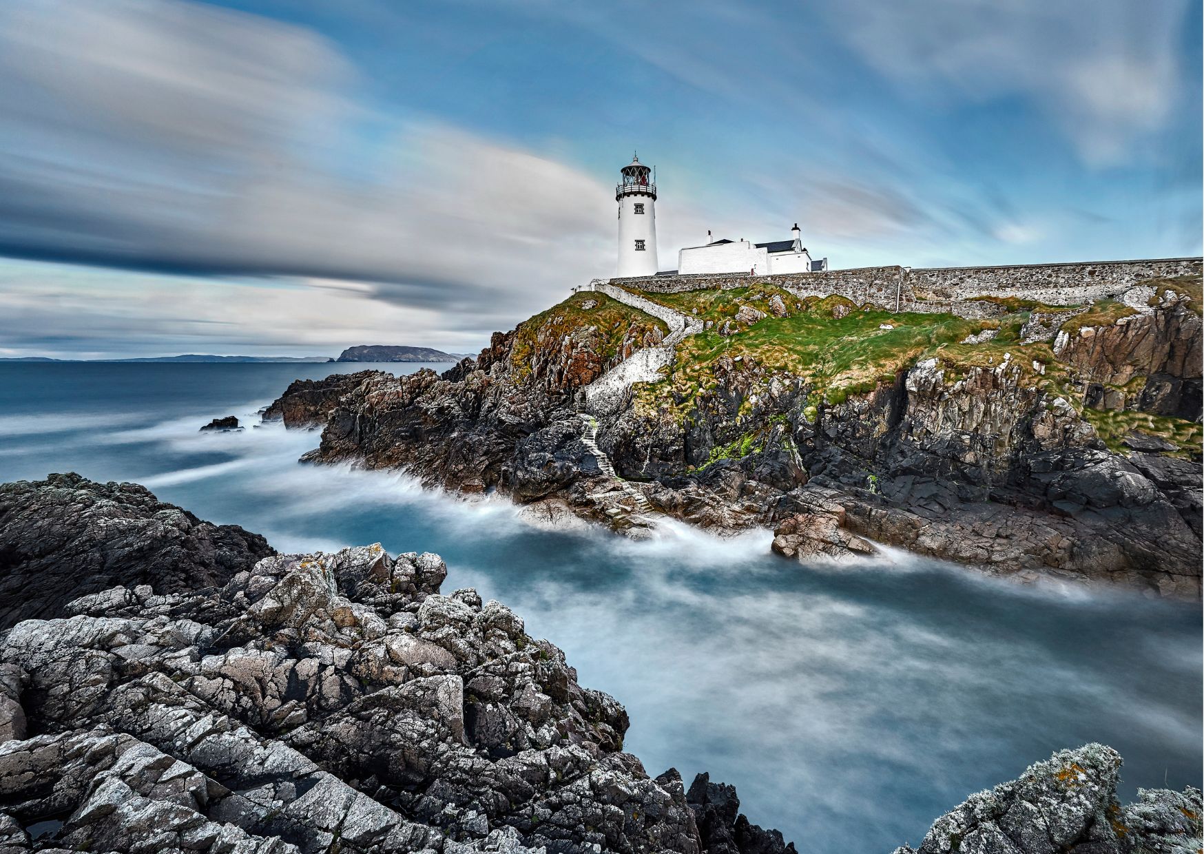 Lighthouse perched on a rocky cliff in Donegal, Ireland, overlooking the Atlantic Ocean under clear skies.