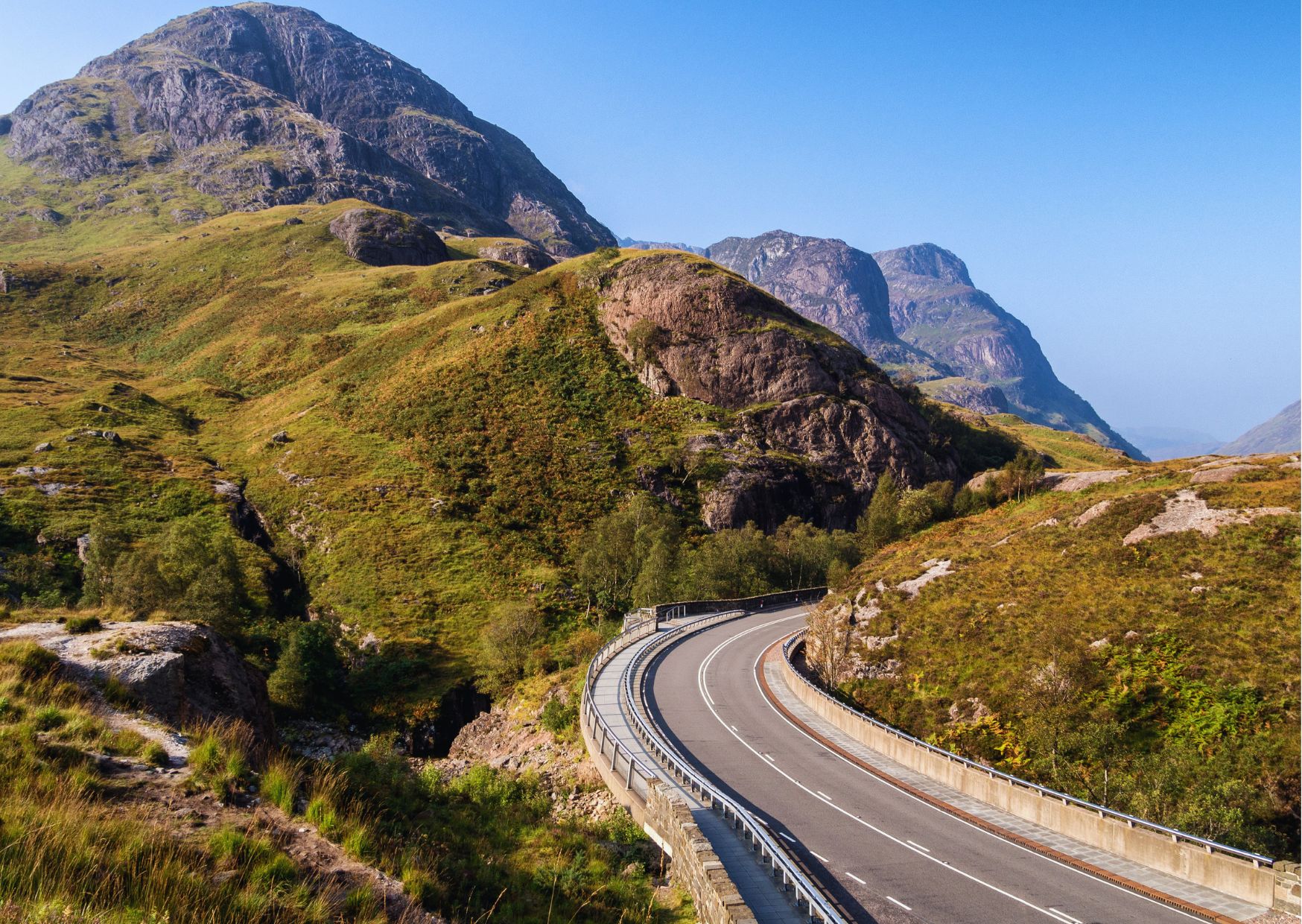Winding road through the dramatic Glencoe valley in Scotland, flanked by towering mountains and open moorland.