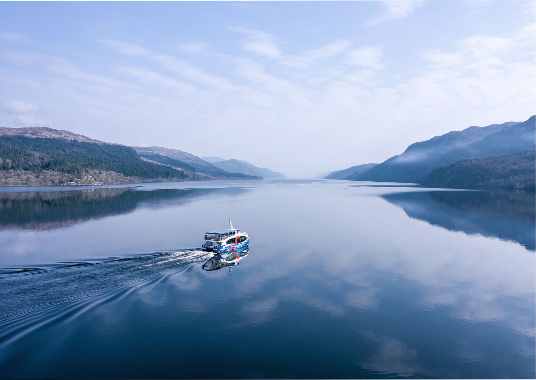 Boat cruising on the calm waters of Loch Ness beneath clear blue skies and surrounded by green hills