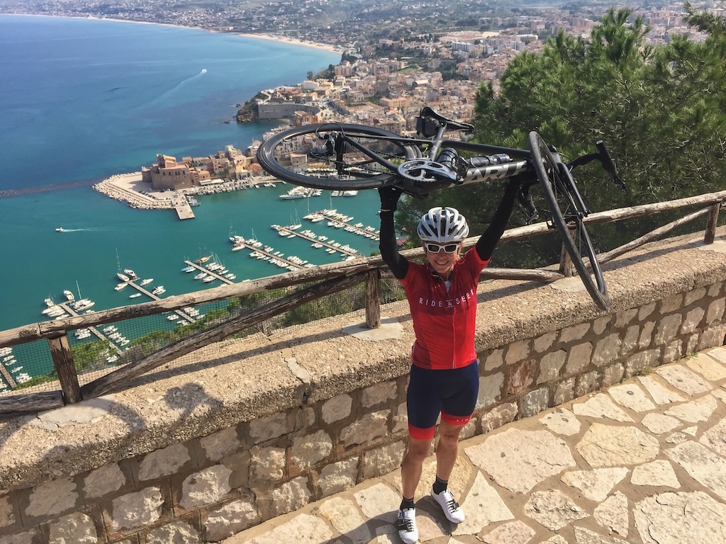 Cyclist holding a road bike overhead at a scenic coastal overlook with harbor and town below.